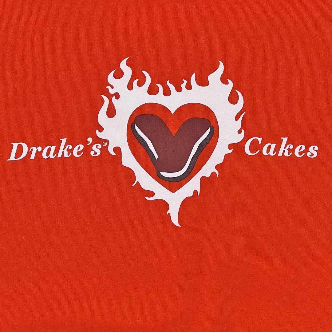 Close-up of the logo on Drake’s Devil Dog Delight orange hoodie, featuring the white text 'Drake’s Cakes' on either side of a stylized heart. The heart, outlined in white, contains a graphic of a Devil Dog pastry with a chocolate exterior and a cream-filled center, set against a fiery, flame-like backdrop.