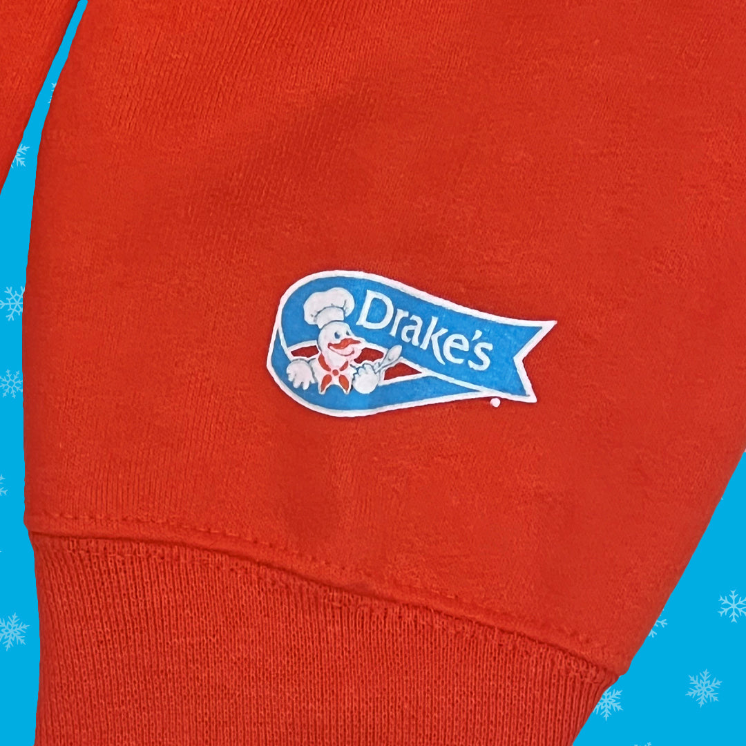 Detail of the sleeve on a Devil Dog Delight Hoodie orange hoodie featuring the Drake's logo. The logo includes an illustration of Webster, the Drake's Cake mascot, a smiling chef with a white hat, within a blue banner. The background is a vibrant orange fabric, and the image is framed by a blue edge with white snowflake accents.