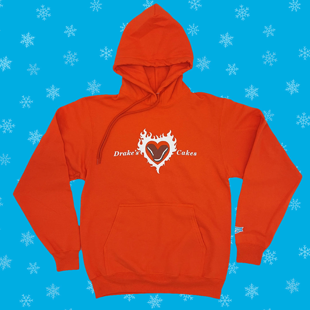 Bright orange hoodie featuring the Drake's Cakes logo with a heart-shaped Devil Dog design, set against a blue background sprinkled with white snowflakes. The hoodie has a full-length zipper, a front pouch pocket, and is presented in a flat lay fashion.