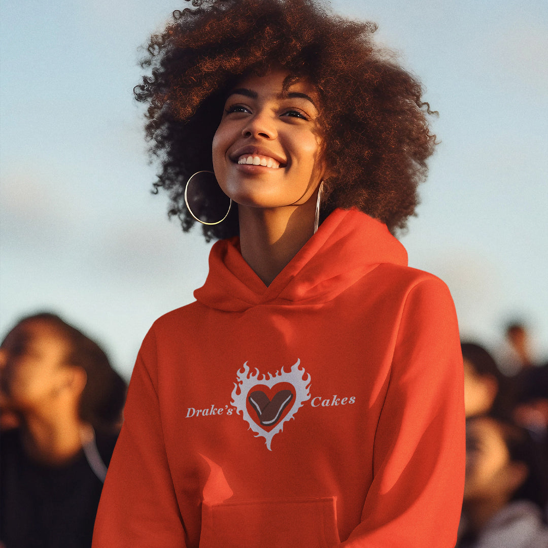 A woman with a radiant smile and curly hair wears a vibrant orange Devil Dog Delight hoodie featuring Drake's Cakes text with a heart-shaped Devil Dog design on the chest. She is wearing large hoop earrings, and the background captures a softly lit sky at dusk with a crowd of people, suggesting an outdoor event or gathering.