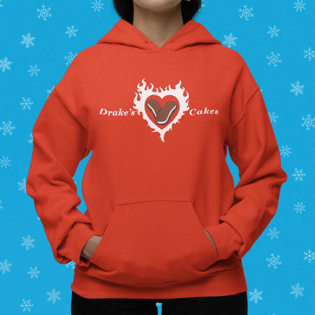 A person stands wearing the Drake’s Devil Dog Delight Hoodie in bold orange, hands casually placed in the front pouch pocket. The hoodie is emblazoned with the distinctive Drake’s Cakes text around a heart-shaped center featuring a Devil Dog pastry, all set against a vivid blue background decorated with white snowflakes