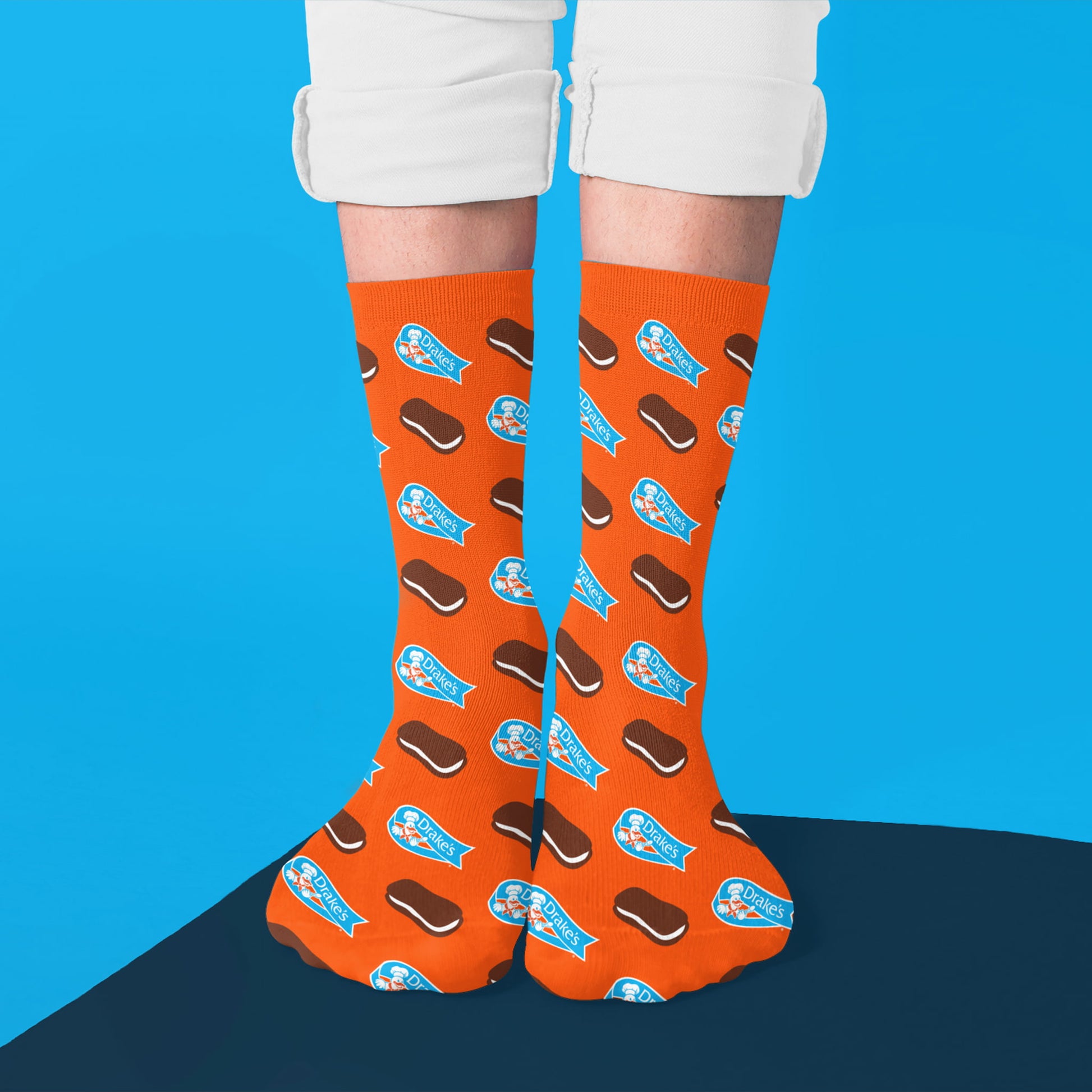 A pair of orange Devil Dogs® socks being worn, featuring a fun pattern of mini Devil Dogs snack cakes and blue Drake's® logo banners. The snack cakes are brown with a cream filling, while the logos and cakes are evenly scattered across the socks. The socks are styled with cuffed white pants and shown against a two-tone blue background.