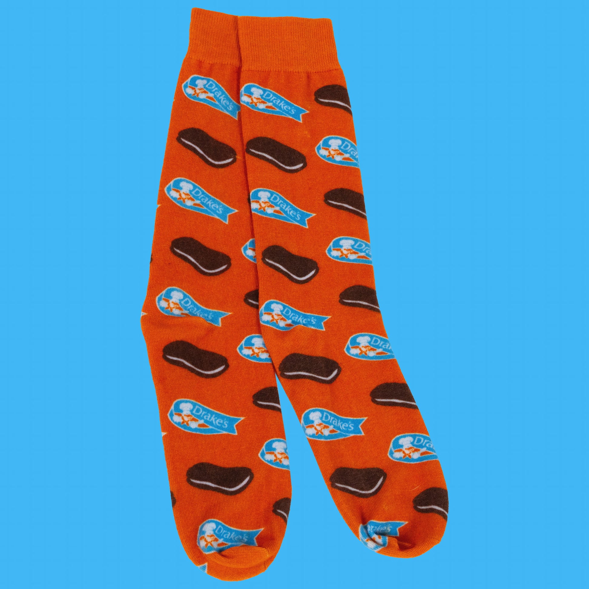 A pair of bright orange Devil Dogs® socks featuring a playful pattern of mini Devil Dogs snack cakes and the Drake's® logo. The snack cakes are brown with a cream filling, and the blue Drake's® logo banners are scattered evenly across the socks. The socks have a solid orange cuff, heel, and toe area. The background is a solid light blue color.