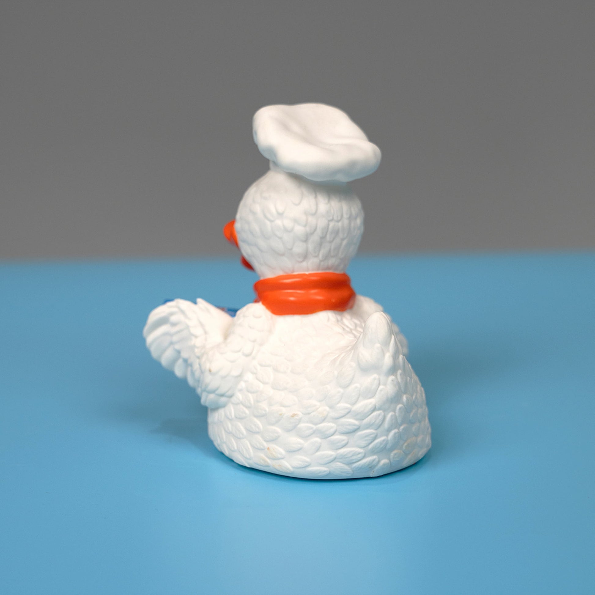 A rear view of the Webster the Duck figurine, the mascot of Drake's Cakes. The rubber duck figurine depicts a white duck with textured feathers, dressed in a chef's hat and a red bandana. Webster is holding a wooden spoon and a box of Devil Dogs. The background is a light blue surface.