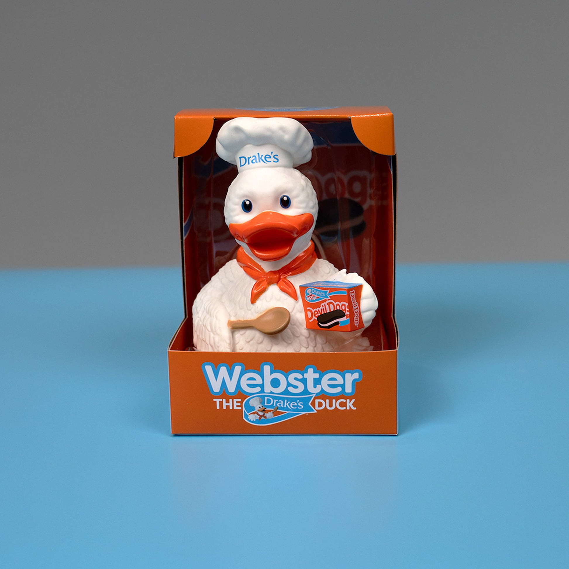 A rubber figurine of Webster the Duck, the mascot of Drake's Cakes, displayed in its packaging. Webster is a cheerful white duck dressed in a chef's hat with "Drake's" written on it and an orange bandana. He is holding a wooden spoon and a box of Devil Dogs. The packaging is orange and blue, with "Webster the Drake's Duck" written on the front. The figurine is set against a light blue background.