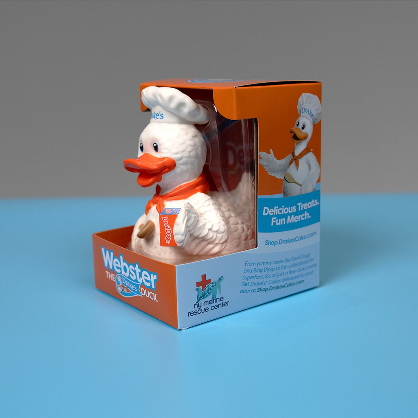 A side view of the Webster the Duck figurine, the mascot of Drake's Cakes, displayed in its packaging. Webster is a white duck dressed in a chef's hat with "Drake's" written on it and an orange bandana. He is holding a wooden spoon and a box of Devil Dogs. The packaging is orange and blue, with "Webster the Drake's Duck" written on the front. The side of the packaging features information about supporting the NY Marine Rescue Center and promotes Drake's Cakes merchandise. 