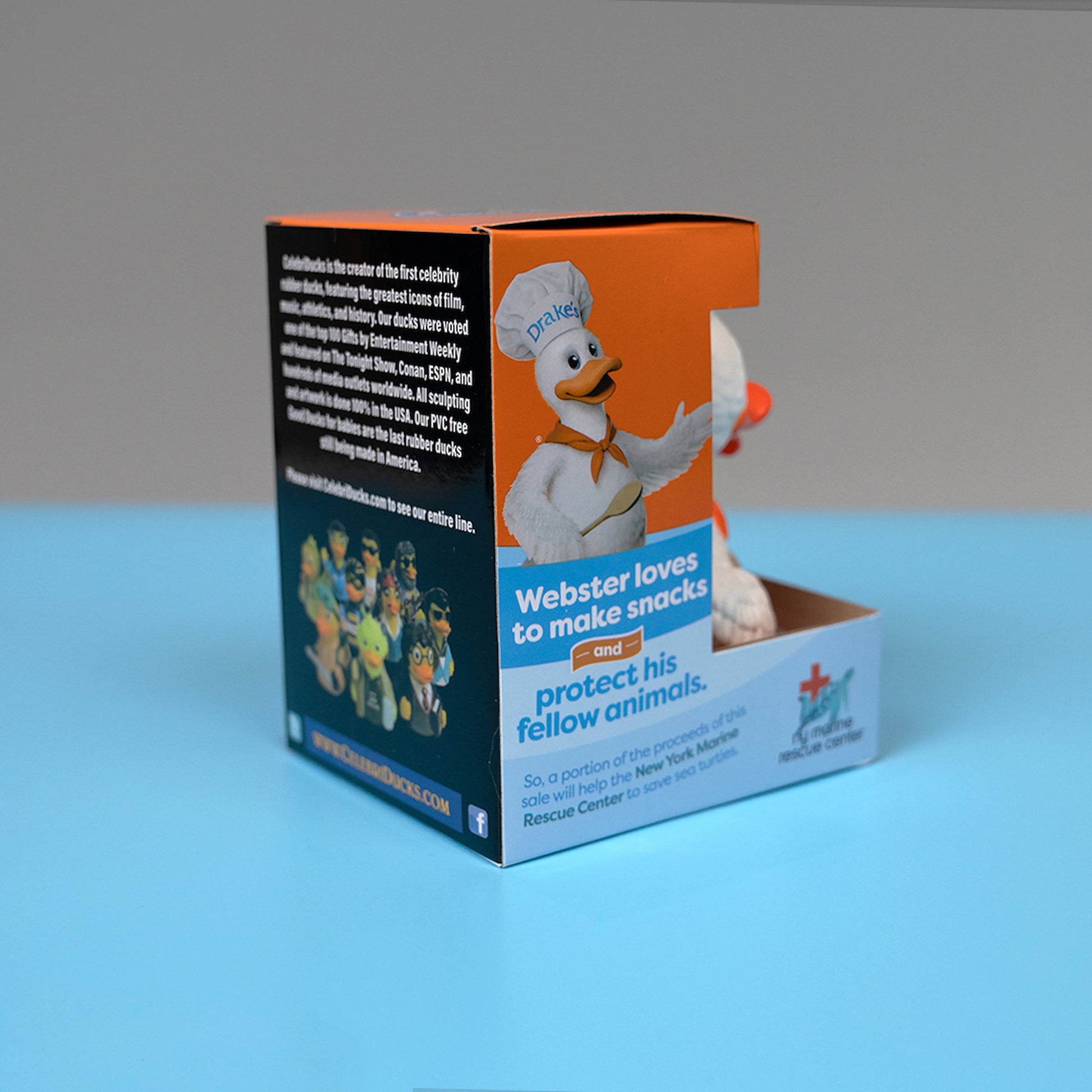 A side view of the packaging for the Webster the Duck figurine. The box features Webster, a white duck dressed in a chef's hat and orange bandana, holding a wooden spoon. The side of the packaging includes an image of Webster with the text "Webster loves to make snacks and protect his fellow animals," highlighting that a portion of the proceeds supports the New York Marine Rescue Center. The back of the box provides information about CelebriDucks. 