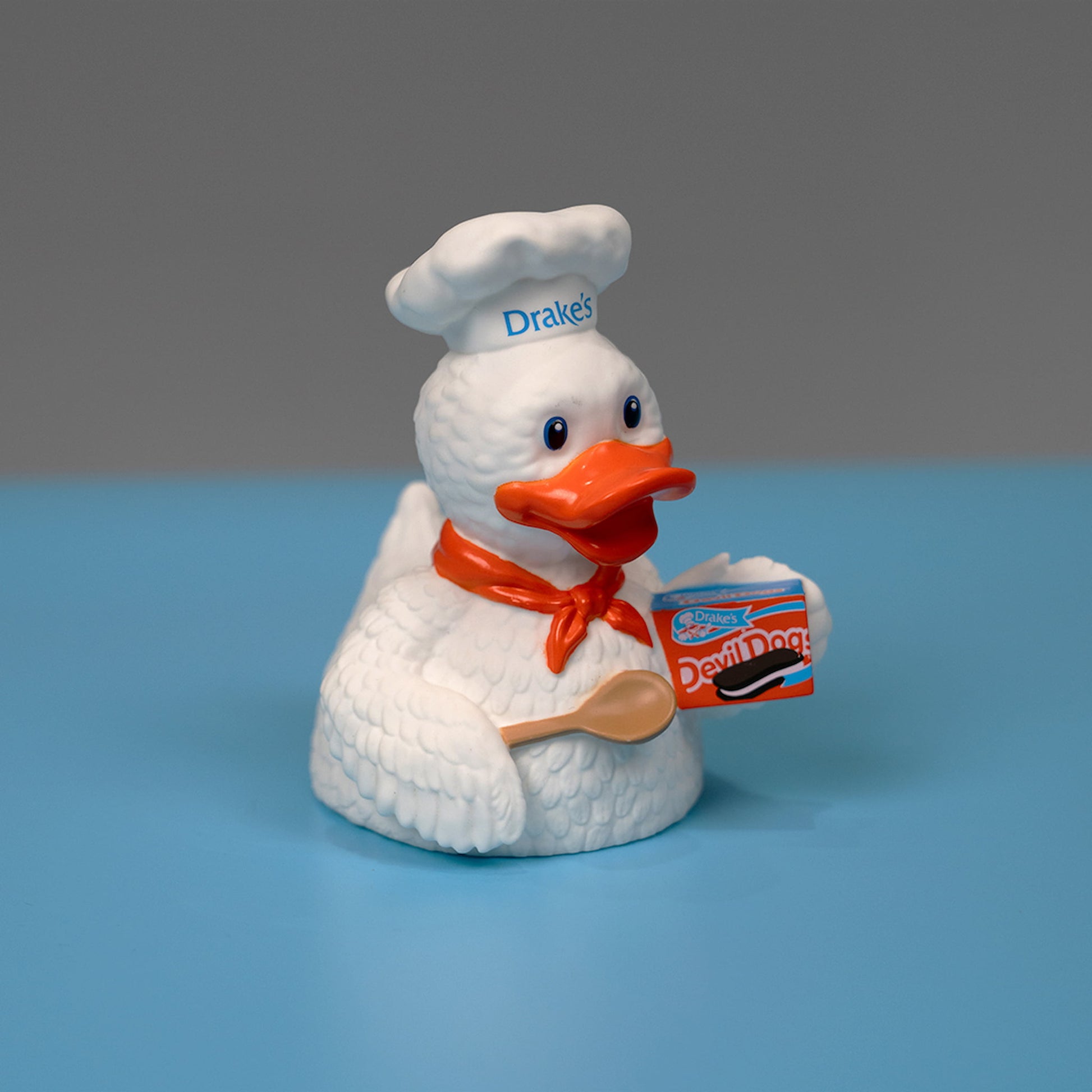 A rubber figurine of Webster the Duck, the mascot of Drake's Cakes. Webster is depicted as a cheerful white duck dressed in a chef's hat with "Drake's" written on it, and a red bandana. He is holding a wooden spoon and a box of Devil Dogs. The figurine is set against a light blue background.