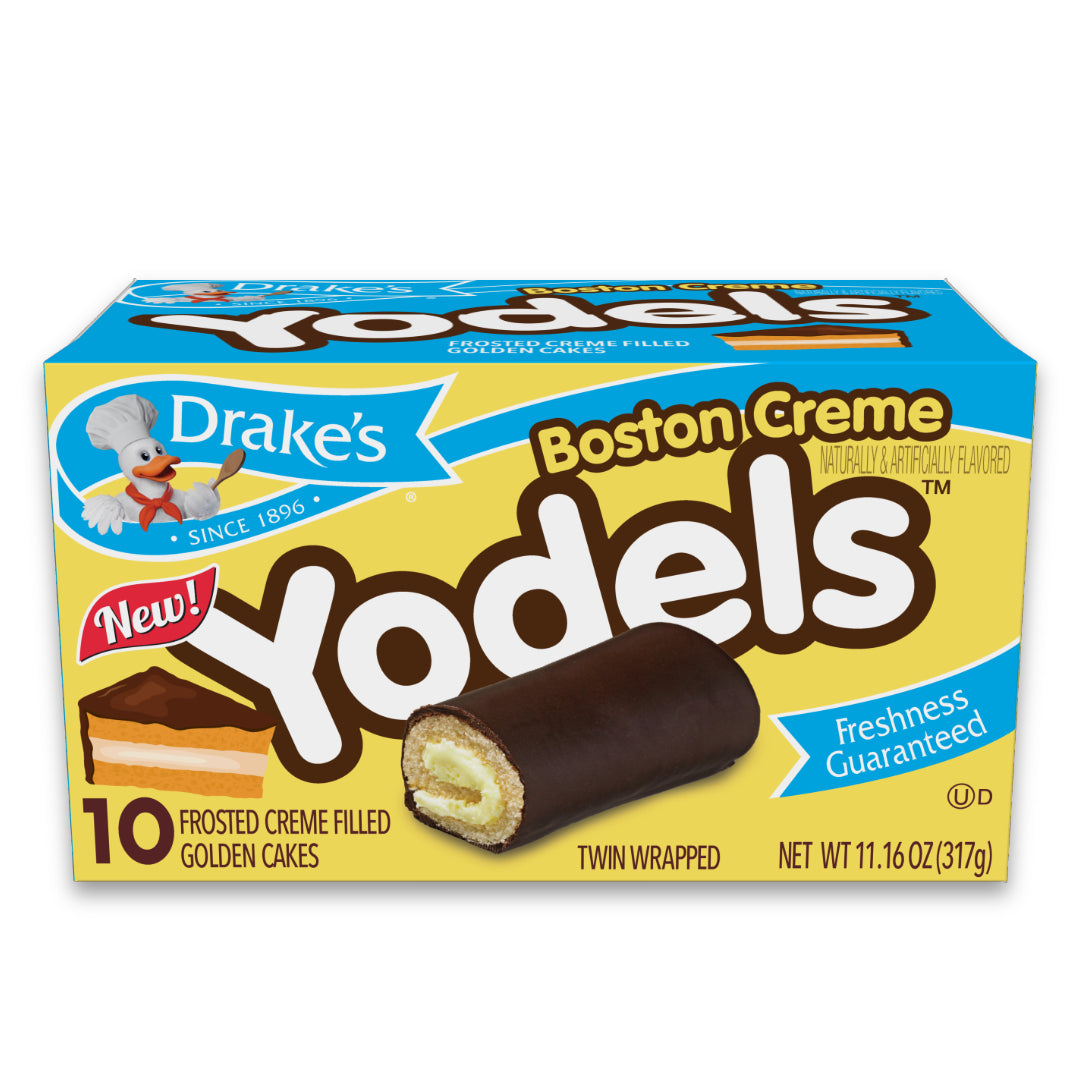 Package of Drake's Boston Creme Yodels. The box is bright blue with a large 'Yodels' text in brown and a 'New!' label in red. Below, 'Boston Creme' is written in a yellow bubble font. The Drake's logo, featuring Webster the duck, is on the upper left with 'Since 1896' beneath it. The package boasts '10 Frosted Creme Filled Golden Cakes', 'Twin Wrapped', and a 'Freshness Guaranteed' seal. Net weight is listed as 11.16 oz (317g)