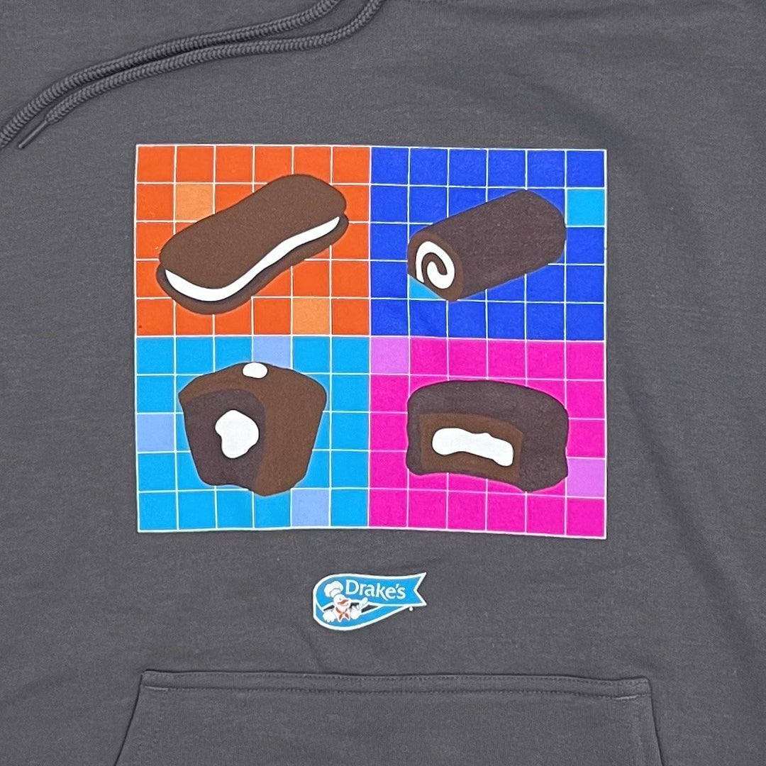 Close-up of the Drake’s Snack-Tastic Hoodie's chest area, featuring a colorful graphic of assorted Drake’s Cakes snacks on a checkered background of orange, blue, and pink squares. Prominently displayed snacks include a Devil Dog, a Ring Ding, Yankee Doodle and a Yodel. A small Drake's logo is visible above the hoodie's large front pocket, set on a grey fabric backdrop.