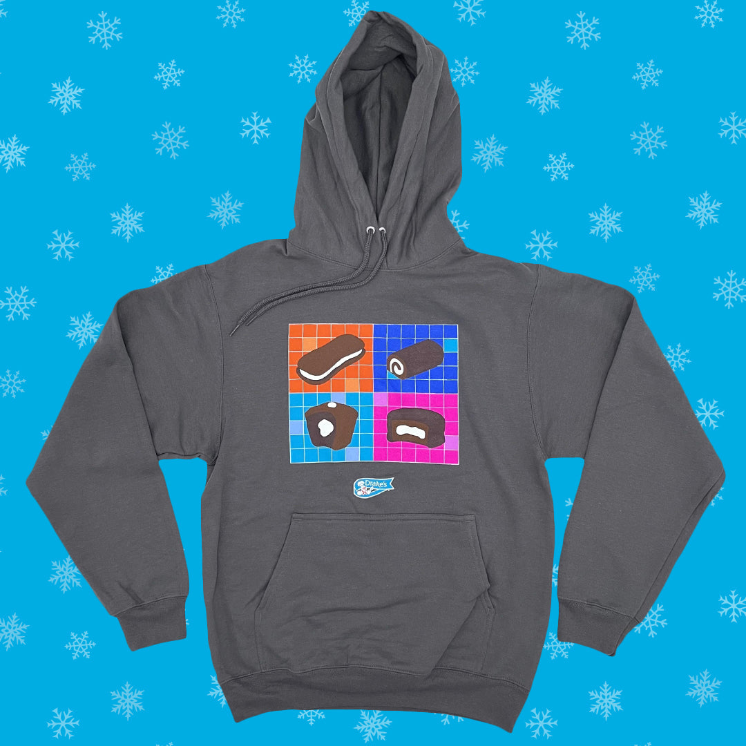 A grey Drake’s Snack-Tastic Hoodie laid flat against a blue background with white snowflakes. The front showcases a vibrant, colorful grid pattern with illustrations of Drake’s iconic snacks, including Devil Dogs, Ring Dings, Yankee Doodles and Yodels. The hoodie has a full hood with drawstrings and a front pouch pocket, featuring the Drake’s logo located above the pocket.