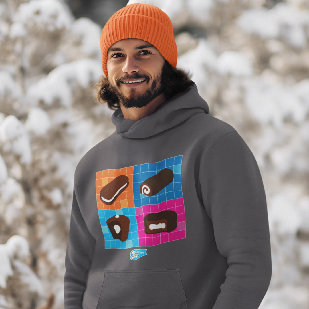 A man with a warm smile wearing a grey Drake’s Snack-Tastic Hoodie and an orange beanie poses against a winter backdrop with snow-covered trees. The hoodie features a colorful, playful grid with images of classic Drake’s snacks on the chest and a small Drake’s logo above the pouch pocket.
